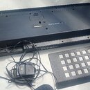 Fotografie zachycuje spodní stranu černého plastového elektronického klávesu s nápisem YAMAHA. Vedle nástroje je položen černý napájecí adaptér s kab…