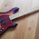 Elektrická basová kytara s trupem v přechodovém lakování od žluté přes oranžovou po tmavě hnědou. Nástroj má červený vzorovaný pickguard a světle hně…