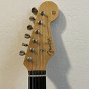 Fotografie zachycuje hlavu elektrické kytary z přírodního světlého dřeva s chromovanými ladičkami. Na povrchu je černý nápis „Fender STRATOCASTER“ a…