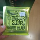 Zelené balení kytarových strun s nápisy ERNIE BALL, REGULAR SLINKY a CUSTOM GAUGE® 2221. Na obalu je uveden seznam tlouštěk strun (10, 13, 17, 26, 36…