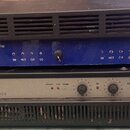 Dva rackové výkonové zesilovače naskládané na sebe. Horní zařízení je modré s černým horním krytem a nápisy PROFESSIONAL POWER AMPLIFIER, logo AG aku…