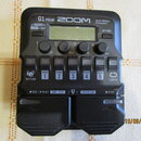 Černý multi-efektový procesor s nápisy G1 FOUR, ZOOM a Multi-Effects Processor. Zařízení má malý displej, čtyři otočné ovladače s popisky Lo, Mid, Hi…