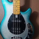 Elektrická basová kytara s modrým třpytivým povrchem a světle hnědým hmatníkem. Nástroj má černý pickguard, na kterém jsou umístěny dva snímače s náp…