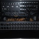 Fotografie zachycuje rack s audio vybavením v černém transportním kufru. V horní části je umístěno zařízení s řadou ovládacích prvků a zapojenými kab…