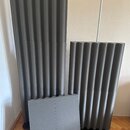 Tři kusy šedého akustického absorbentu z pěnového materiálu. Dva kusy mají vlnovitý povrch a jeden kus je plochý. Všechny jsou umístěny na dřevěné po…