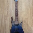 Elektrická kytara s modrým přechodovým lakem a viditnou strukturou dřeva na hranách. Na hlavě hmatníku je bílý nápis Ibanez. Nástroj má dva humbucker…