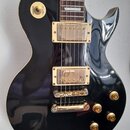 Černá elektrická kytara s gold hardwarem a hmatníkem z tmavého dřeva. Na přepínači tónu jsou nápisy RHYTHM a TREBLE. Kytara má dva humbuckery s kryty…