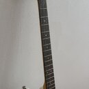 Bílá elektrická kytara s hnědým hmatníkem a světle dřevěným krčkem. Na těle je vidět tmavý pickguard a jeden ladicí kolík na hlavě. Povrch nástroje v…