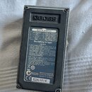 Fotografie zachycuje spodní stranu efekt pedálu značky BOSS v šedé barvě. Na spodní straně je nalepený černý štítek s tabulkou BOSS AC ADAPTOR, infor…