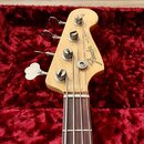 Hlava elektrické basy z přírodního dřeva s tmavým hmatníkem. Na hlavě je vidět logo Fender a nápis JAZZ BASS ELECTRIC BASS. Nástroj má čtyři kovové l…