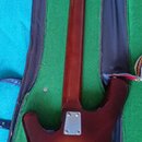 Fotografie zachycuje zadní stranu elektrické kytary v obdélníkovém kufru s zeleným vnitřkem. Kytara má hnědý sunburst povrch a dřevěný krk. Na zadní…