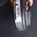 Detailní pohled na zadní část černého kulatého zařízení z plastu. Na boku jsou dva konektory, jeden s červeným prstencem. Na bílé nálepce je uveden t…