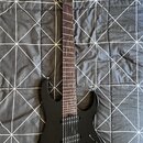 Černá elektrická kytara s tmavým hmatníkem a dvěma humbuckery. Na hlavě je bílý nápis Ibanez. Nástroj vykazuje známky běžného používání, jsou vidět d…