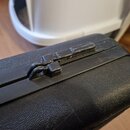 Detailní pohled na horní hranu černého kufru z texturovaného materiálu. Je vidět plastový zapínací mechanismus, který vykazuje známky opotřebení a od…