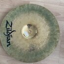 Kovový činel zlatavé barvy s viditelným zeleným oxidem a patinou po okrajích. Na povrchu je černý nápis Zildjian. Povrch vykazuje známky opotřebení a…