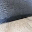 Detailní pohled na spodní část černého reproduktorového boxu s texturovaným povrchem a černou ochrannou tkaninou. Na spodním hraně rámu jsou viditeln…