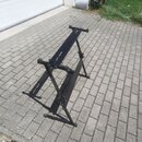 Černý kovový stojan na klávesové nástroje v rozloženém stavu. Stojan má X-tvarovou konstrukci s nastavitelnou výškou a dvěma horními oporami. Na povr…