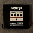 Černá krabice s bílými nápisy ORANGE AMPLIFICATION a TERROR STAMP GUITAR AMPLIFIER PEDAL. Uprostřed je vyobrazen bílý efekt s nápisy ORANGE TERROR ST…