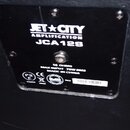 Fotografie zachycuje zadní část černého reproduktoru s připevněným černým plastovým štítkem. Na štítku je bílý nápis JET CITY AMPLIFICATION JCA12S a…