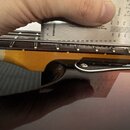 Detailní pohled na hmatník a kobylku strunového nástroje s kovovými strunami. Nad strunami je přiloženo kovové měřidlo s nápisy Millimeters, Inches a…