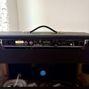 Zadní strana kytarového komba v černém texturovaném potahu s černou rukojetí. Na zadním panelu je nápis TONE MASTER DELUXE REVERB a logo Fender. Je z…