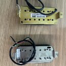 Dvě elektrické snímače kytary s kovovými kryty. Horní snímač má zlatý povrch a bílou nálepu s nápisy „RoHS“, „RoHS“ a „PC117N-XBB 13.01“. Dolní sníma…