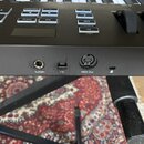 Detail zadní strany černé klávesové nástroje s několika konektory. Jsou zde viditelné nápisy „Sustain“, symbol USB a „MIDI Out“. V horní části jsou t…
