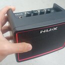 Černý kompaktní kytarový zesilovač s červeným rámečkem kolem přední mřížky. Na mřížce i na horním panelu je bílé logo NUX, přičemž nápis na horní čás…