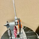 Stříbrný kovový mechanismus pedálu s dlouhou pákou, která má na konci kulatý černý úchop s bílým vrchem. Součástí je pohon s řetězem a oranžovým ozub…