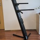 Černý kovový stojan na klávesové nástroje s dvěma vodorovnými rameny. Stojan má šikmý hlavní sloup s integrovanou rukojetí pro přenos a křížovou zákl…