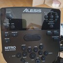 Černý elektronický modul k bubnům s nápisem ALESIS v horní části a označením NITRO DRUM MODULE vlevo dole. Zařízení obsahuje displej, několik ovládac…