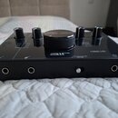 Černý audio rozhraní s lesklým povrchem. Na horní straně jsou čtyři menší otočné ovladače a jeden velký centrální knoflík. Na povrchu je bílý nápis "…
