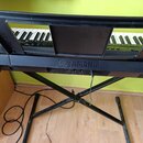 Černý digitální klavír umístěný na černém X-stojanu. Na přední hraně korpusu je bílý nápis YAMAHA s doprovodným logem. Pod nápisem jsou vidět konekto…