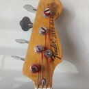 Fotografie zachycuje hlavu basové kytary z přírodního dřeva s четыřmi kovovými ladičkami. Na dřevě je černý nápis Fender Jazz Bass a pod ním drobný t…