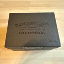 Černá čtvercová krabice s nápisy. Na horní straně je uveden text BLACK COUNTRY CUSTOMS, HANDCRAFTED IN THE UK a LOUDPEDAL. V pravém dolním rohu je po…