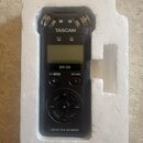 Černý digitální záznamový zařízení v bílém polystyrenovém obalu. Na přístroji je nápis TASCAM, označení modelu DR-05 a v dolní části text LINEAR PCM…