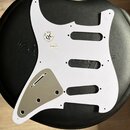 Bílý plastový pickguard pro elektrickou kytaru s šedým krytem snímače. Na povrchu je kulatá nálepka s nápisem PT PSE QC INSPECTION a malý bílý štítek…