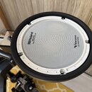 Kruhový elektronický bicí pad s bílou úderovou plochou a černým gumovým okrajem. Na povrchu jsou nápisy „Roland“ s logem a text „MESH PAD Reg. U.S. P…