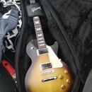 Elektrická kytara s trupem v barevnosti sunburst a tmavým hmatníkem, umístěná v černém polotvrdém pouzdře. Na černém popruhu je bílý nápis Gibson. Tr…