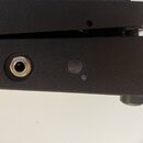 Detailní pohled na černý, matný povrch hudebního zařízení. V levé části je vidět kovový vstupní konektor typu jack. Vedle něj se nachází malý kulatý…