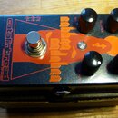 Černý efekt pedál s oranžovou grafikou a nápisy. Na levé straně je svisle napsáno "catalinbread" a pod tím menším písmem "MECHANISMS OF MUSIC", upros…