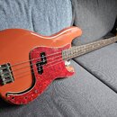Oranžová elektrická basová kytara s červeným vzorovaným pickguardem. Na hlavě hmatníku je viditelné logo Fender. Nástroj má čtyři struny a chromovaný…