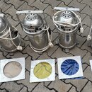 Čtyři stříbrné kovové reflektory s rukojetím a bílými napájecími kabely. Před nimi jsou umístěny čtyři čtvercové kovové rámy s barevnými filtry v ods…