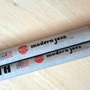 Dvě dřevěné bubenecké paličky s natürálním povrchem. Na obou je červený kruh s bílým nápisem MJC4 a černý text modern jazz COLLECTION. V levé části j…