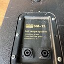 Zadní strana černého reproduktoru s texturovaným povrchem. Na černé tabulce jsou vidět logo DAP AUDIO a označení SM-12, dále nápisy full range system…