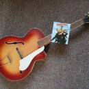 Akustická kytara s vyvedeným korpusem, bílým pickguardem a povrchem v červeno-žlutém sunburst odstínu. Na hmatníku je položen papírový lístek s nápis…