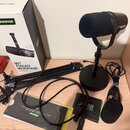 Sada obsahuje černý mikrofon s windsockem na stolním stativu, černé rameno mikrofonu a červený zvukový interface s nápisem Focusrite. Vedle je přibal…