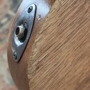 Detailní pohled na zadní část dřevěného hudebního nástroje s kovovým přez covertem a jackovým vstupem. Kovový kryt je připevněn dvěma šrouby a vykazu…