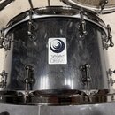 Černý buben s tmavým, mramorovaným povrchem a chromovaným kováním. Uprostřed korpusu je bílá čtvercová nálepka s logem a nápisem „jelen DRUMS“. Povrc…