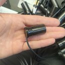 Na dlani je položen malý černý cylindrický mikrofon s připojeným kabelem. Na těle mikrofonu je nápis, který není zcela čitelný. Povrch je matně černý…